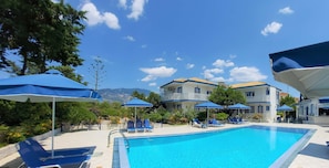 A swimming pool with blue lounge chairs and umbrellas, surrounded by buildings with balconies and a mountainous backdrop.