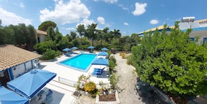 A swimming pool with blue umbrellas, surrounded by greenery and a tiled roof.