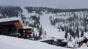 Un edificio moderno con fachada de madera y techo cubierto de nieve, rodeado de un área montañosa boscosa.