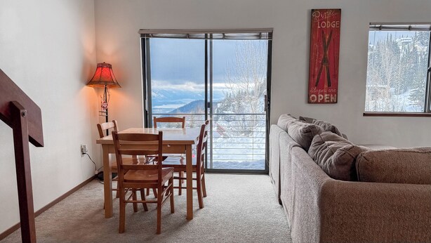 Una sala de estar acogedora con comedor, un amplio ventanal con vistas a montañas nevadas y un letrero que indica que la cabaña está abierta.