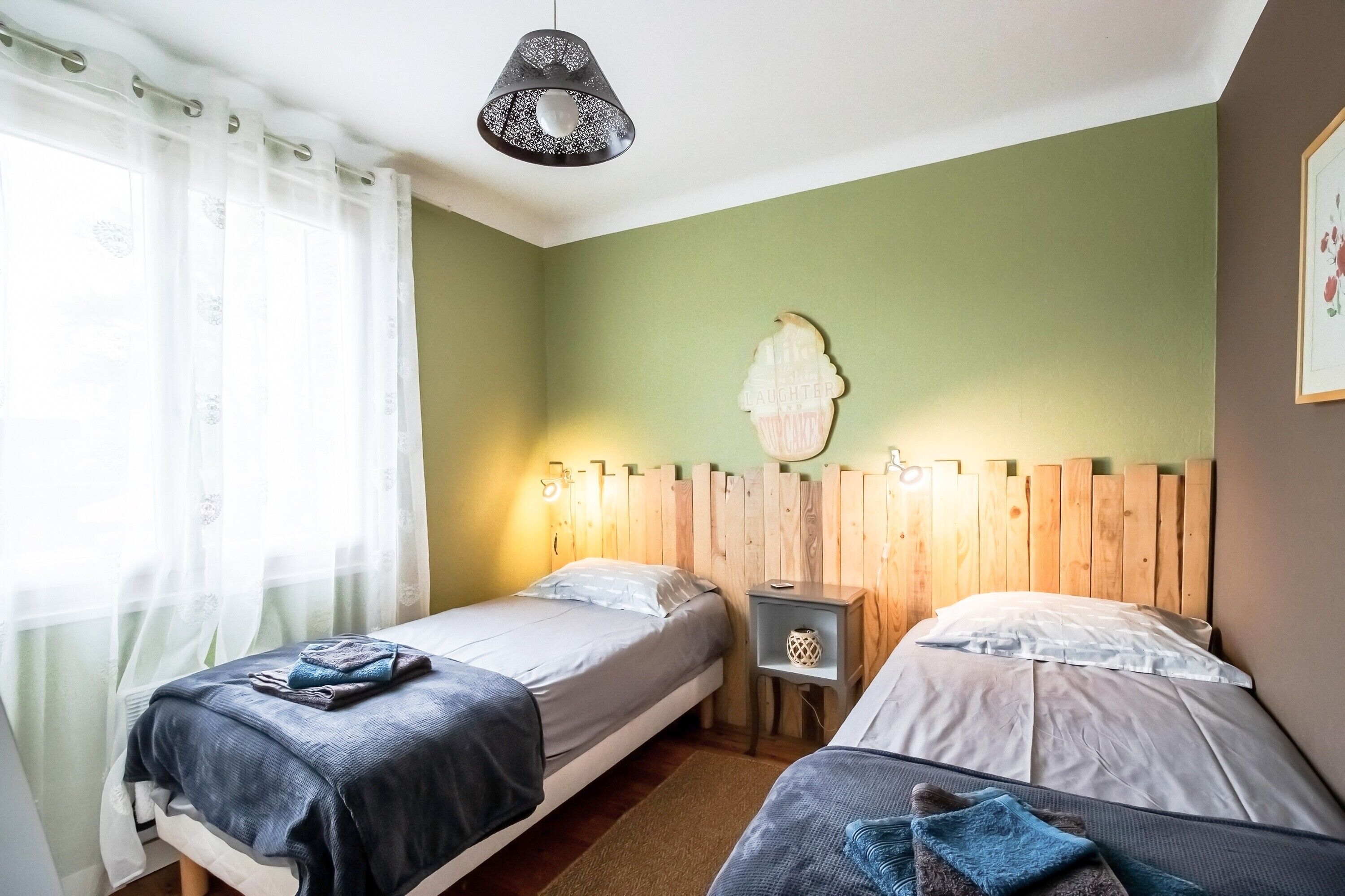 Una habitación con dos camas individuales, cabecera de madera, mesita de noche pequeña, lámpara colgante y una ventana con cortinas traslúcidas.