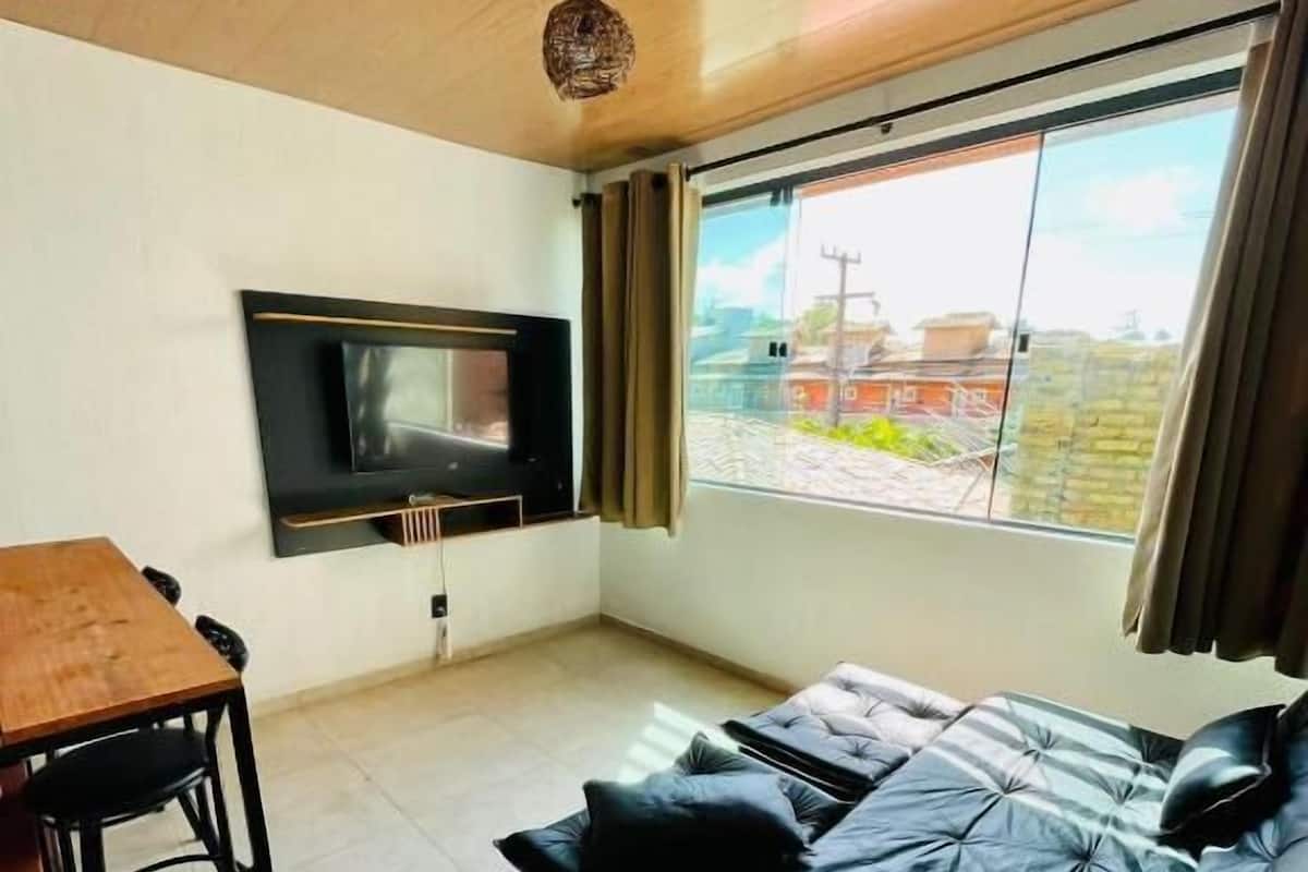 A modern living room with a sofa, a TV mounted on the wall, a dining table, and a large window with curtains.