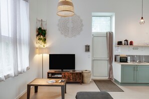 A living room with a flat-screen TV, a wooden coffee table, a hanging plant, a decorative wall piece, and a kitchen area with a microwave and cabinets.