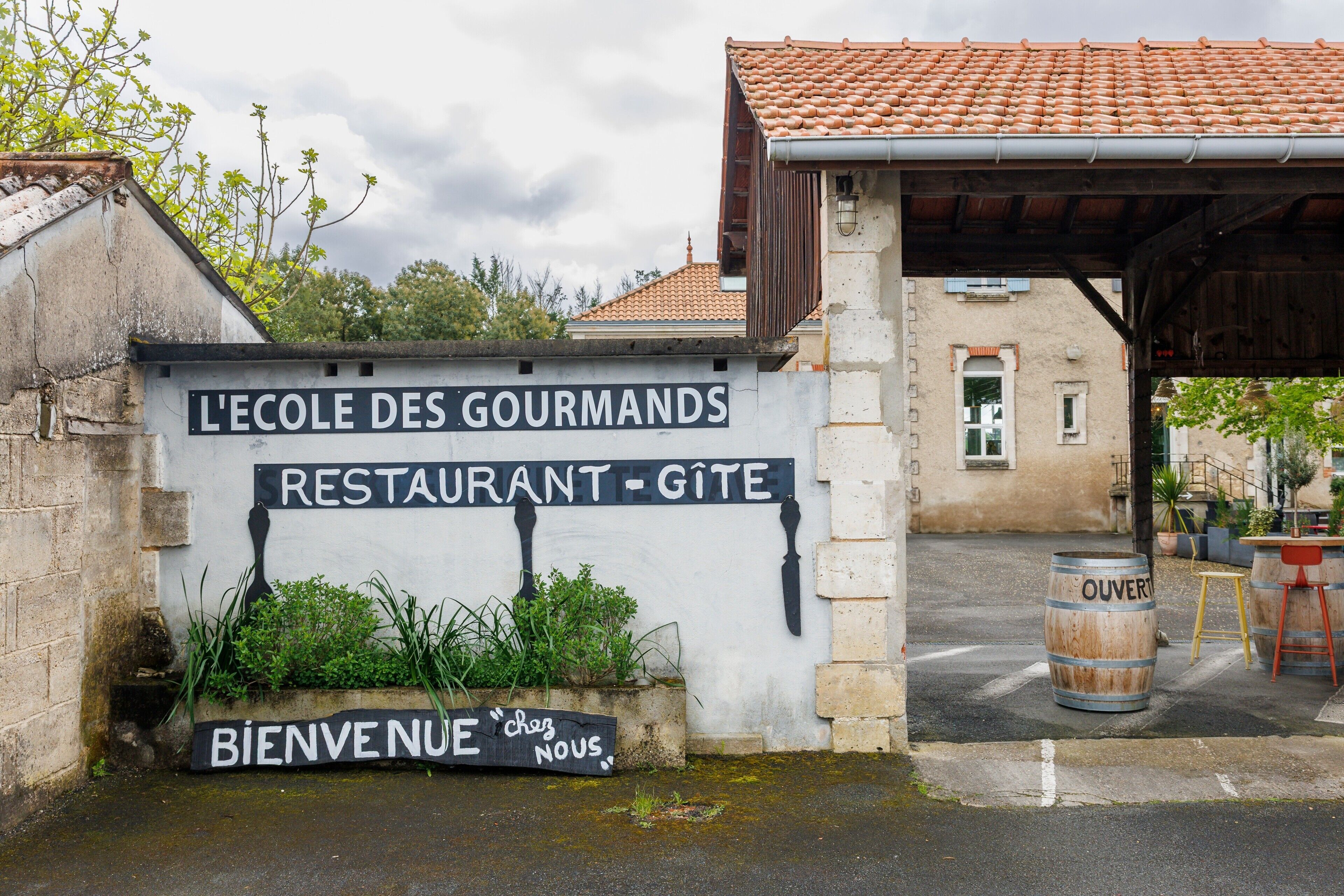 A sign for 'L'Ecole Des Gourmands Restaurant - Gîte' with a welcome sign and a wooden barrel.
