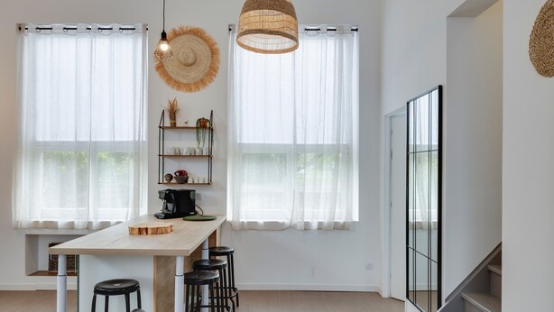 Una cocina moderna con una mesa de madera, taburetes de bar, una cafetera y ventanas amplias con cortinas translúcidas.