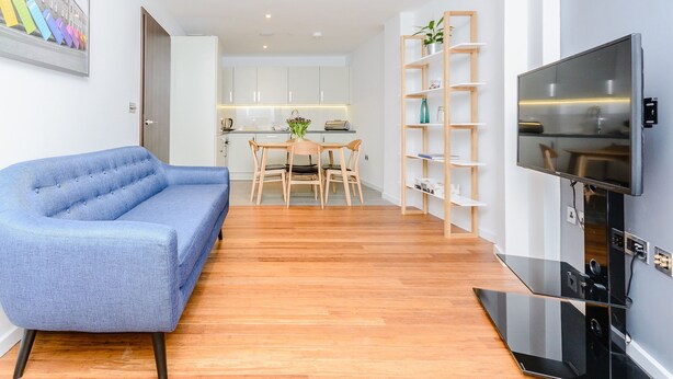 A modern living room with a blue sofa, a dining area with a table and chairs, a kitchen with white cabinets, and a TV mounted on the wall. - Flying Butler Richmond Tersha Street Apartments (Richmond)