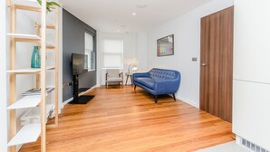 A modern living room with a wooden floor, a blue sofa, a TV, and a bookshelf. - Flying Butler Richmond Tersha Street Apartments (Richmond)