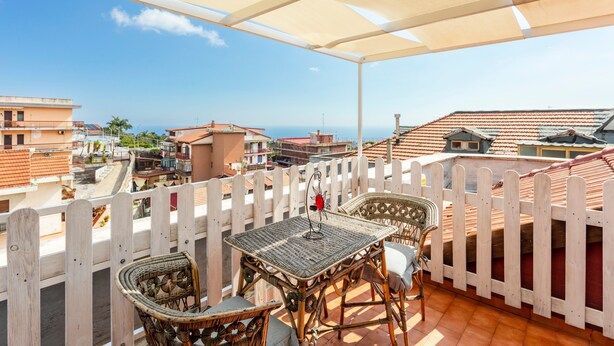 A balcony with wicker furniture, a white picket fence, and a view of the city and sea. - Apartment 'Reale' with Private Terrace, Wi-Fi and Air Conditioning (Gravina di Catania)