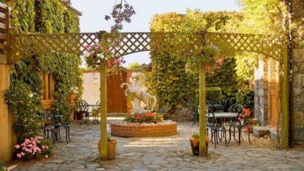 Ein Innenhof mit einem Kiesweg, einer zentralen Fontäne mit einer Statue und einer Pergola, die mit Kletterpflanzen bewachsen ist.