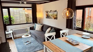 A cozy living room with a grey sofa, white chairs, a wooden table, and a large window with a view of a garden.
