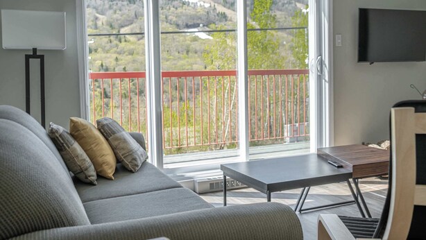 A living room with a sofa, a table, a TV, and a balcony with a view of a hilly landscape.