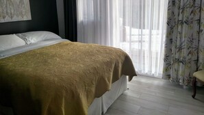 A hotel room with a bed, a chair, a window with sheer curtains, and a wall with a painting.