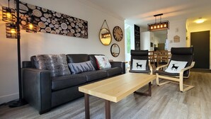 A modern living room with a black leather sofa, two wooden chairs, a wooden coffee table, and a wall-mounted light fixture.