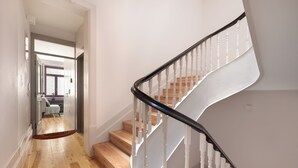 A staircase with white balusters and a black handrail, leading to a landing with a wooden floor and a view of a room with a bed and a window. - Loureiro 140 (Porto)