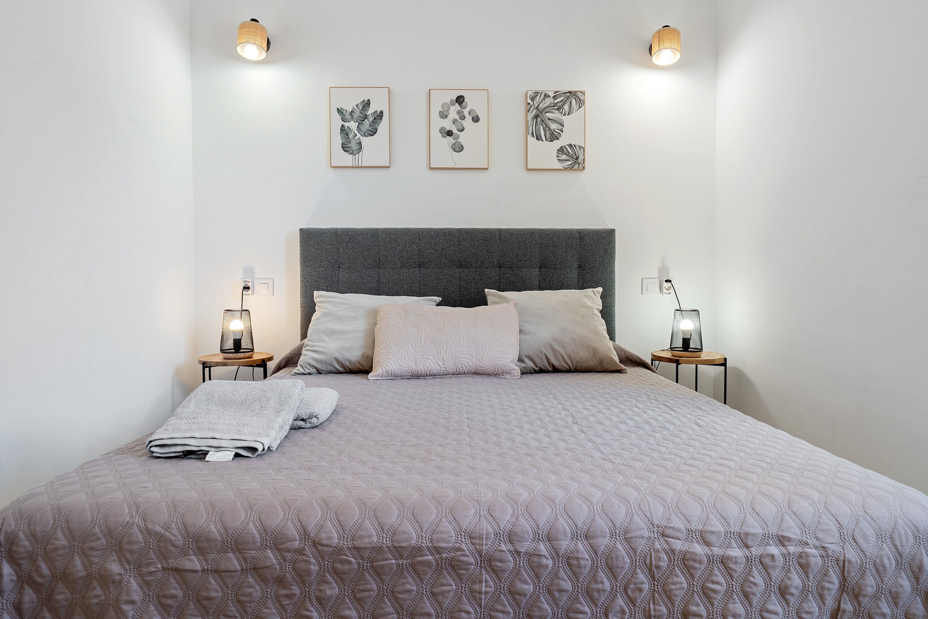 Una cama bien tendida con un edredón acolchado, dos mesitas de noche con lámparas, y tres láminas botánicas enmarcadas en la pared.