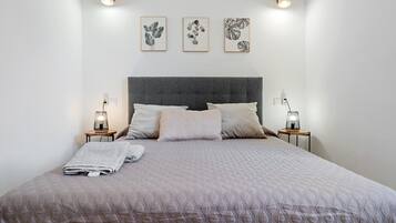 Una cama bien tendida con un edredón acolchado, dos mesitas de noche con lámparas, y tres láminas botánicas enmarcadas en la pared.