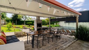 Een terras buiten met een houten tafel en stoelen, een barbecue en een overdekte zithoek.