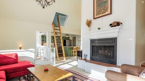 Una sala de estar con chimenea, sofás rojo y beige, una mesa de centro de madera y una cocina al fondo.