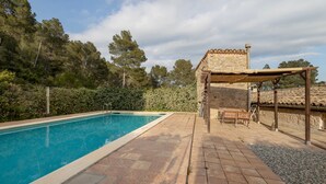 Piscina com deck revestido de azulejos, edifício de pedra com telhado ladrilhado e pérgola de madeira com assentos.
