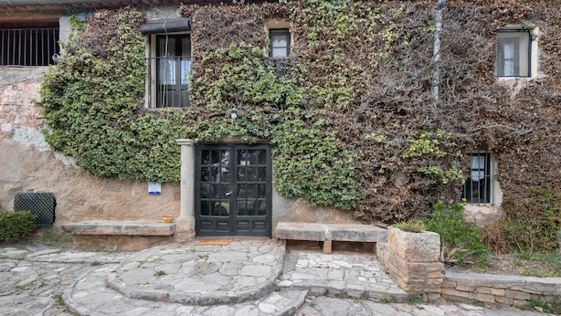 Pátio com pavimento de pedra, um elemento circular de pedra central, uma porta preta e um edifício parcialmente coberto por hera.