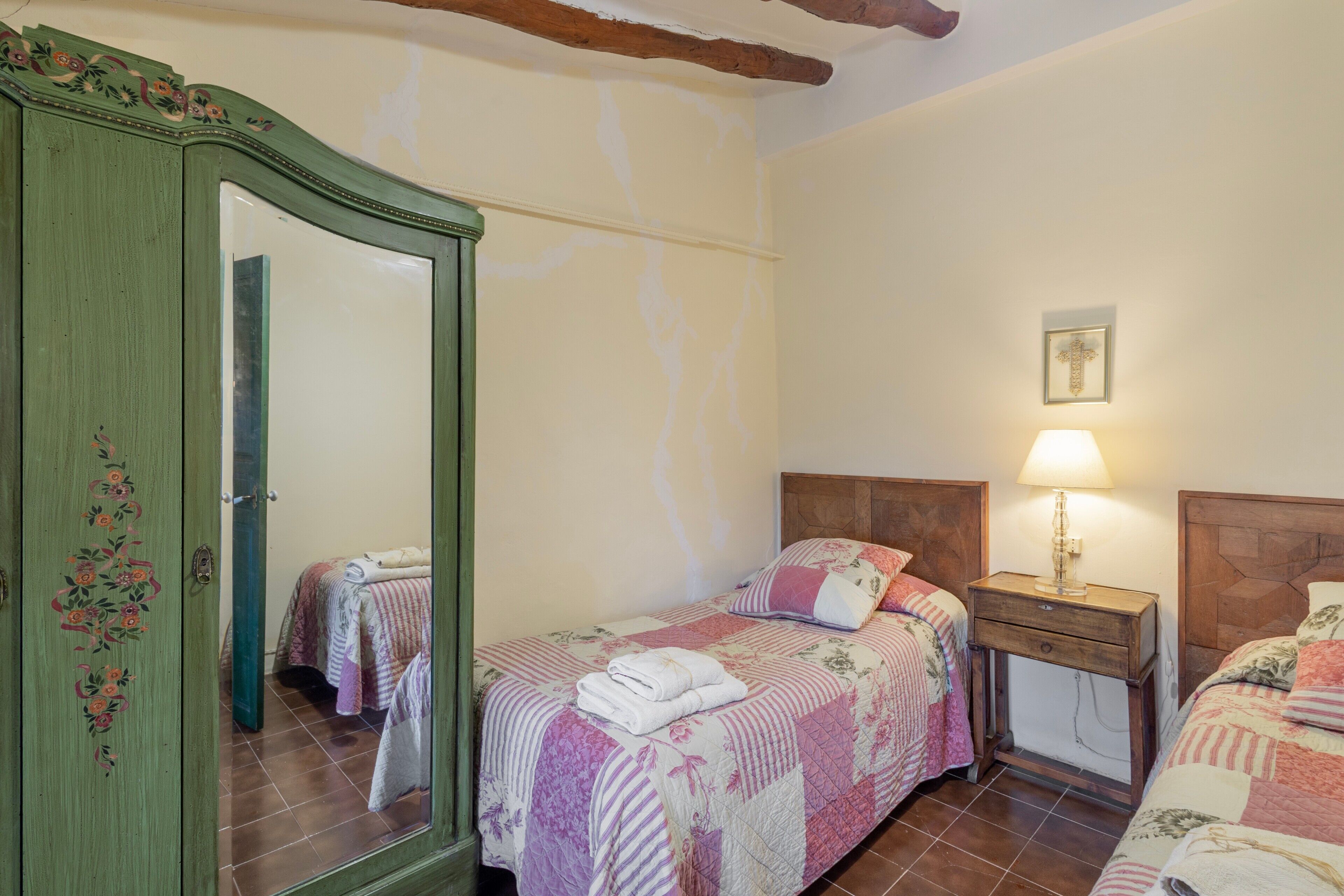 A bedroom with two beds, a wooden wardrobe, a bedside table with a lamp, and a framed picture on the wall.