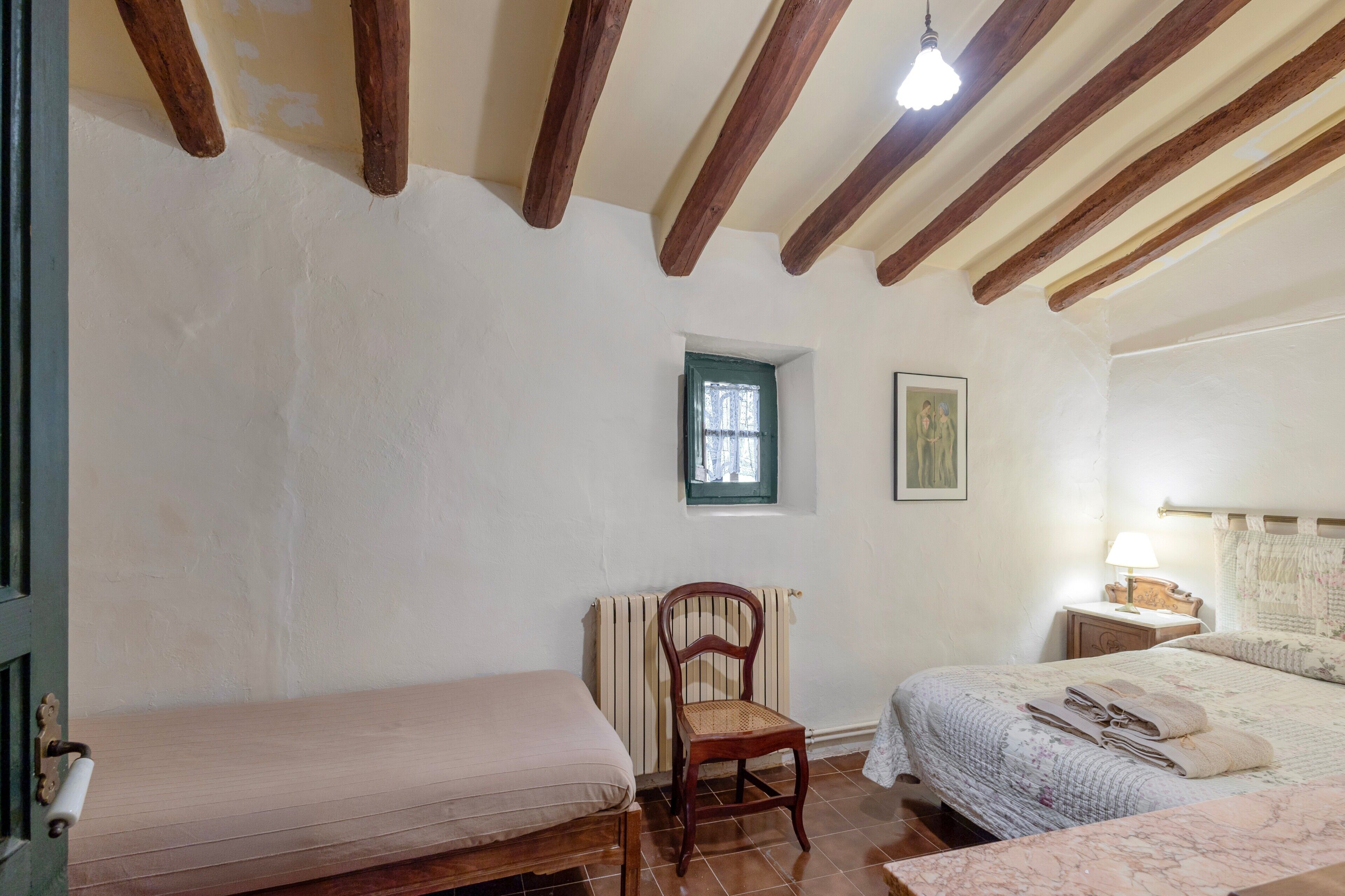 Une chambre avec un lit simple, une chaise, une table de chevet, une petite fenêtre et des poutres en bois au plafond.