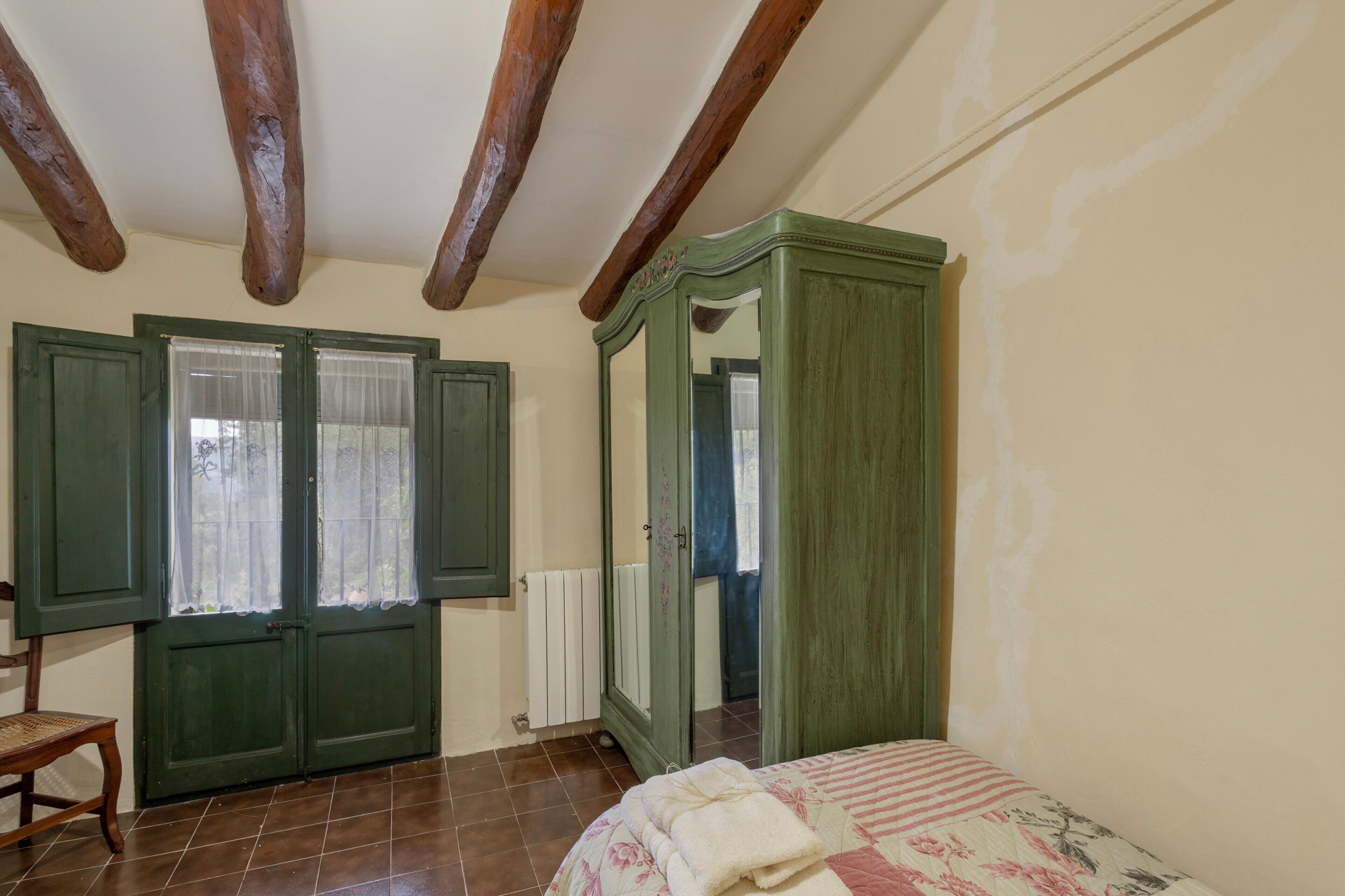Ein Zimmer mit Bett, Holzbalkendecke, einem grünen Kleiderschrank und einem Fenster mit Vorhängen.