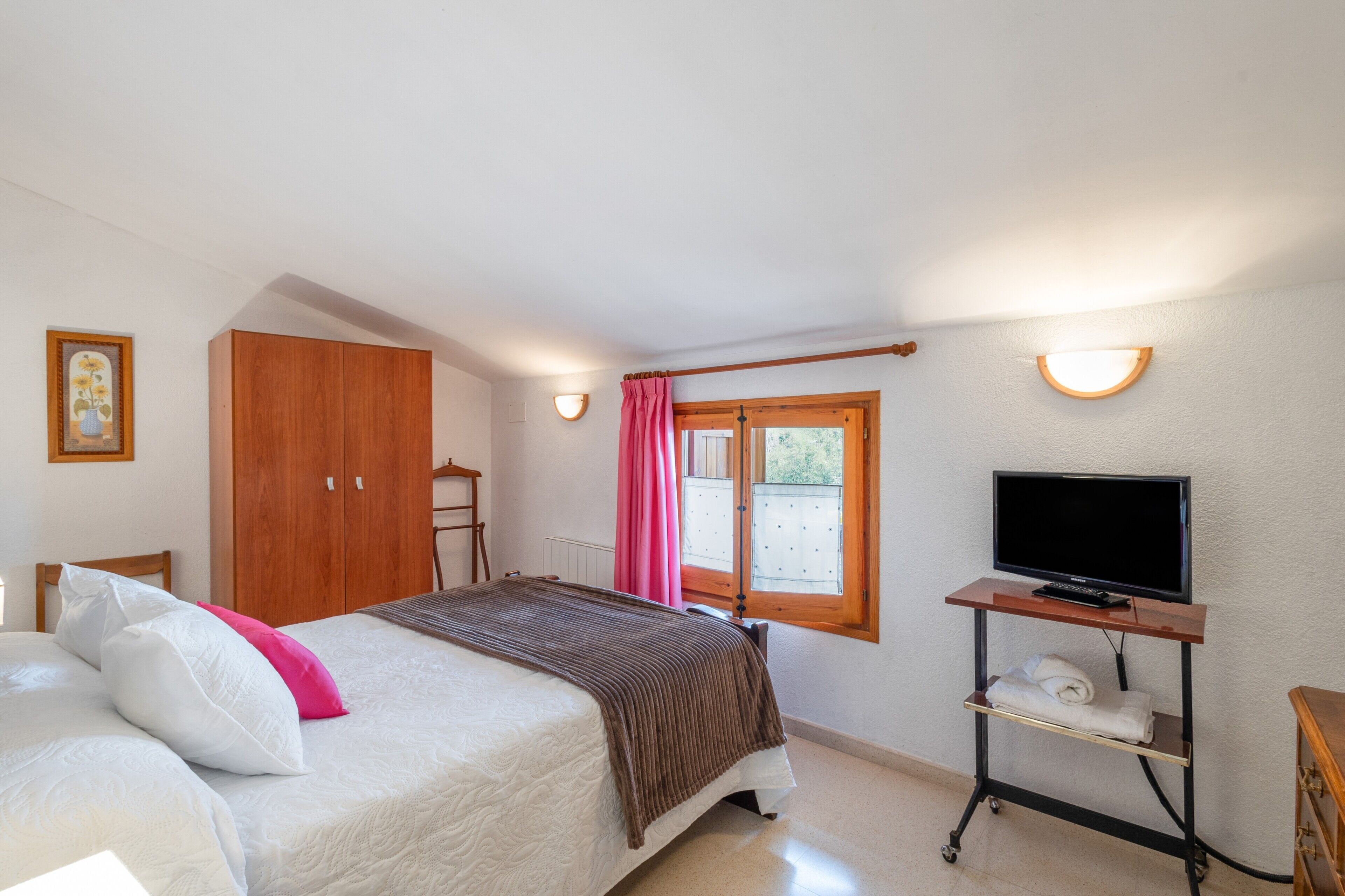 A bedroom with a bed, a TV on a stand, a wardrobe, a window with curtains, and a wall-mounted light.