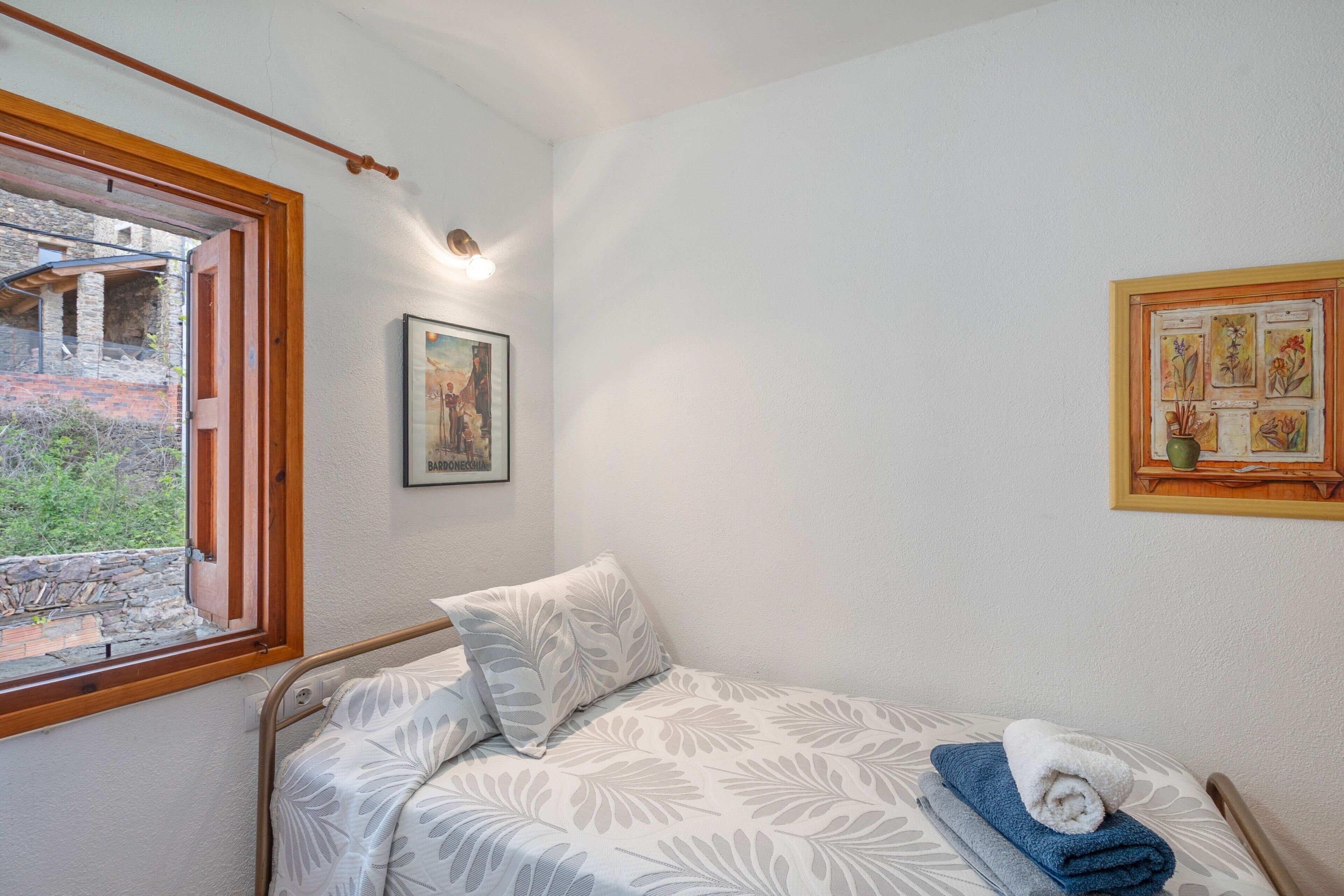 A single bed with a patterned bedspread, a pillow, a rolled towel, a framed picture on the wall, and a window with a view of an outdoor area.