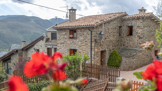 A stone building with a tiled roof, a wooden fence, and a small garden with red flowers.
