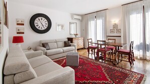 Una sala de estar con un sofá, una mesa de comedor con sillas, una alfombra roja y un reloj de pared.
