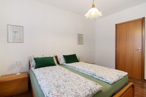 Quarto com duas camas de solteiro, uma mesinha de cabeceira de madeira, um criado-mudo com abajur e uma porta.
