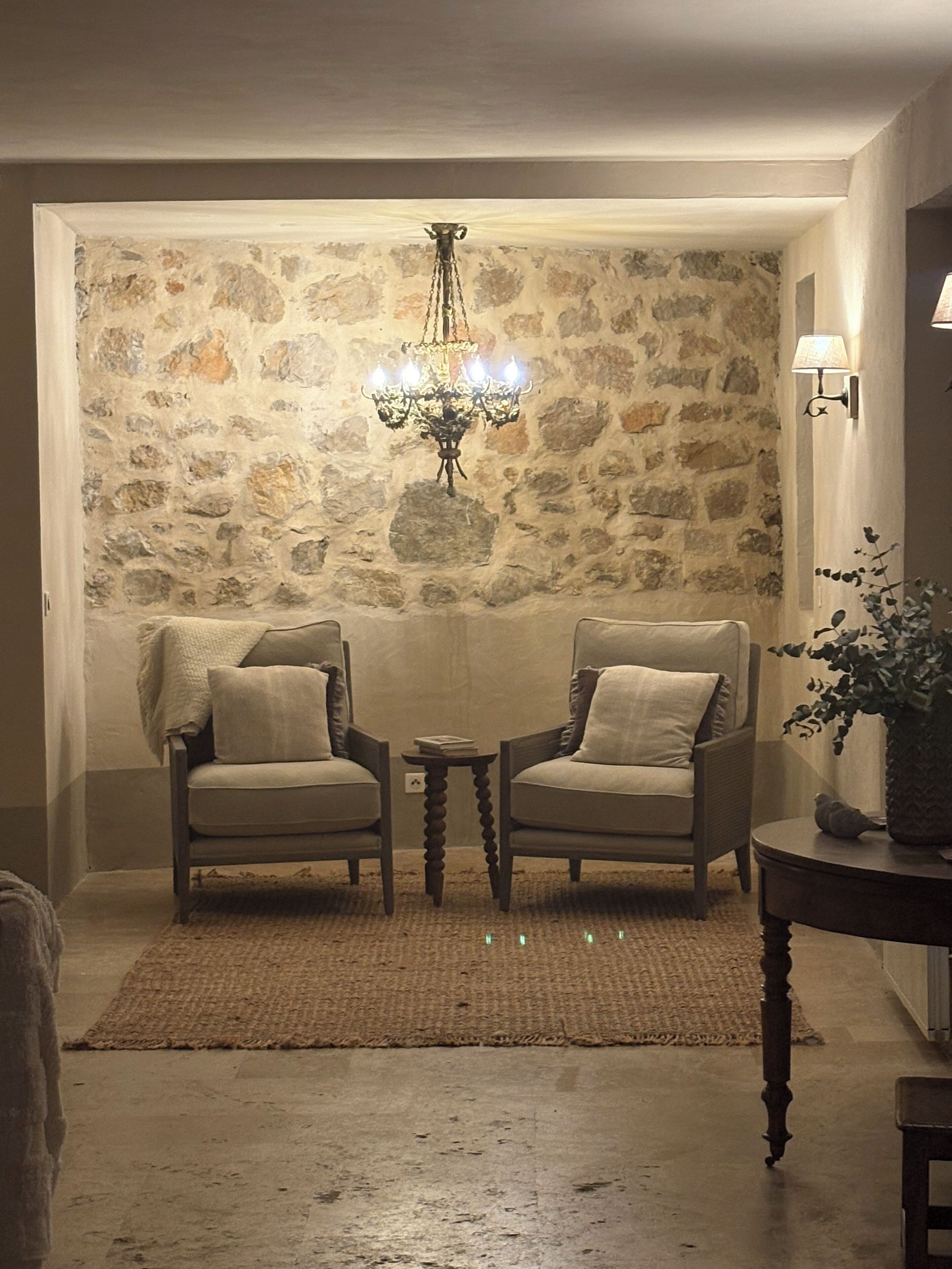 A room with two armchairs, a small table, a chandelier, and a stone wall.