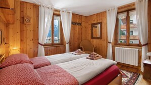 A wooden room with two beds, a chair, a clock, and a window with curtains. - Apartment 'Corti Garden' with Mountain View, Private Garden and Wi-Fi (Campodolcino)