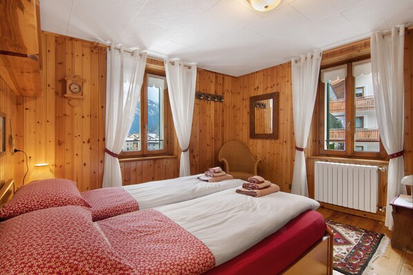 A wooden room with two beds, a chair, a clock, and a window with curtains.