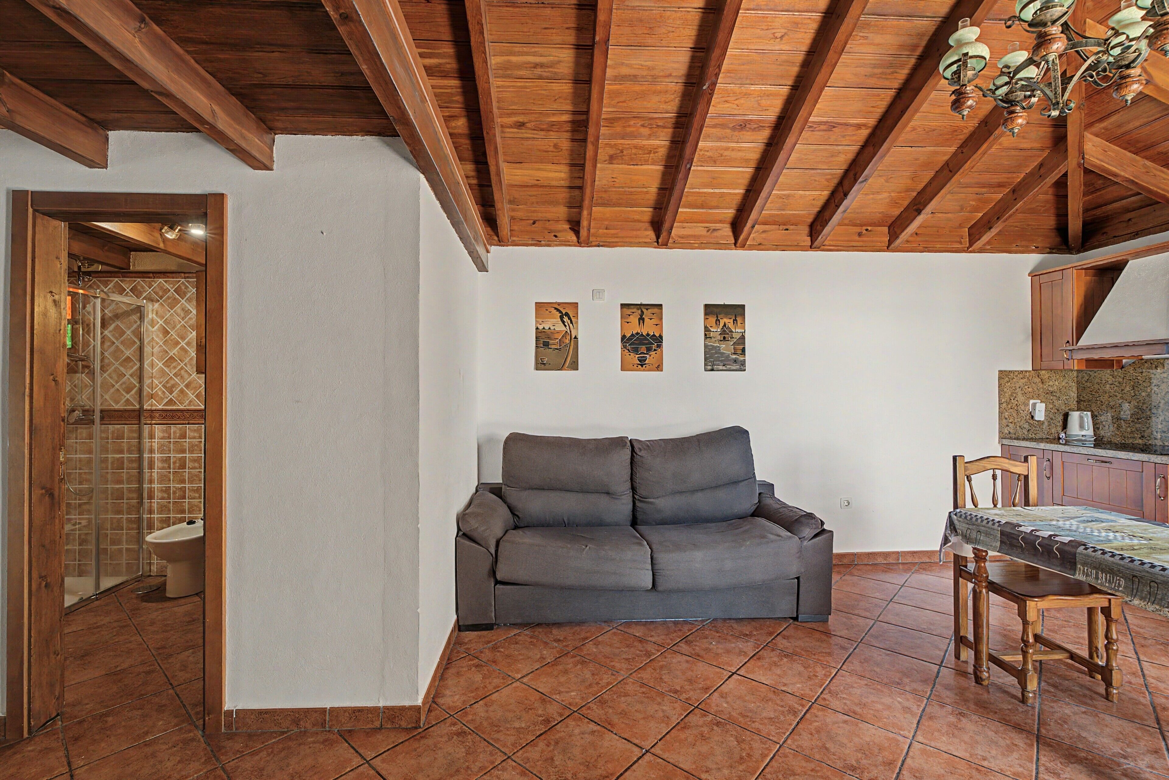 Una habitación con techo de madera, un sofá gris, piso de baldosas y una pequeña mesa con sillas.