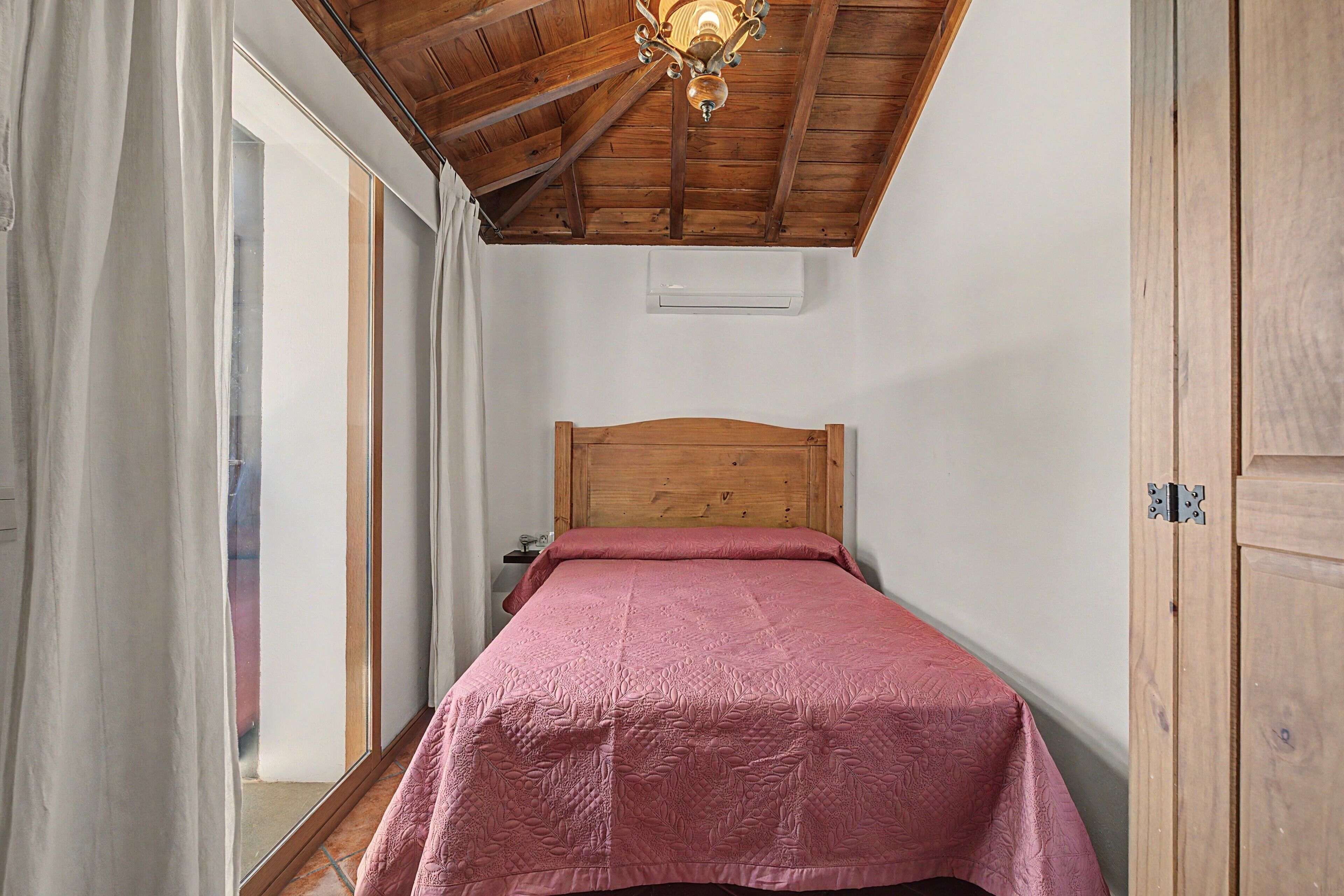Un dormitorio pequeño con techo de madera, una cama individual con colcha rosa, una ventana con cortinas y un armario de madera.