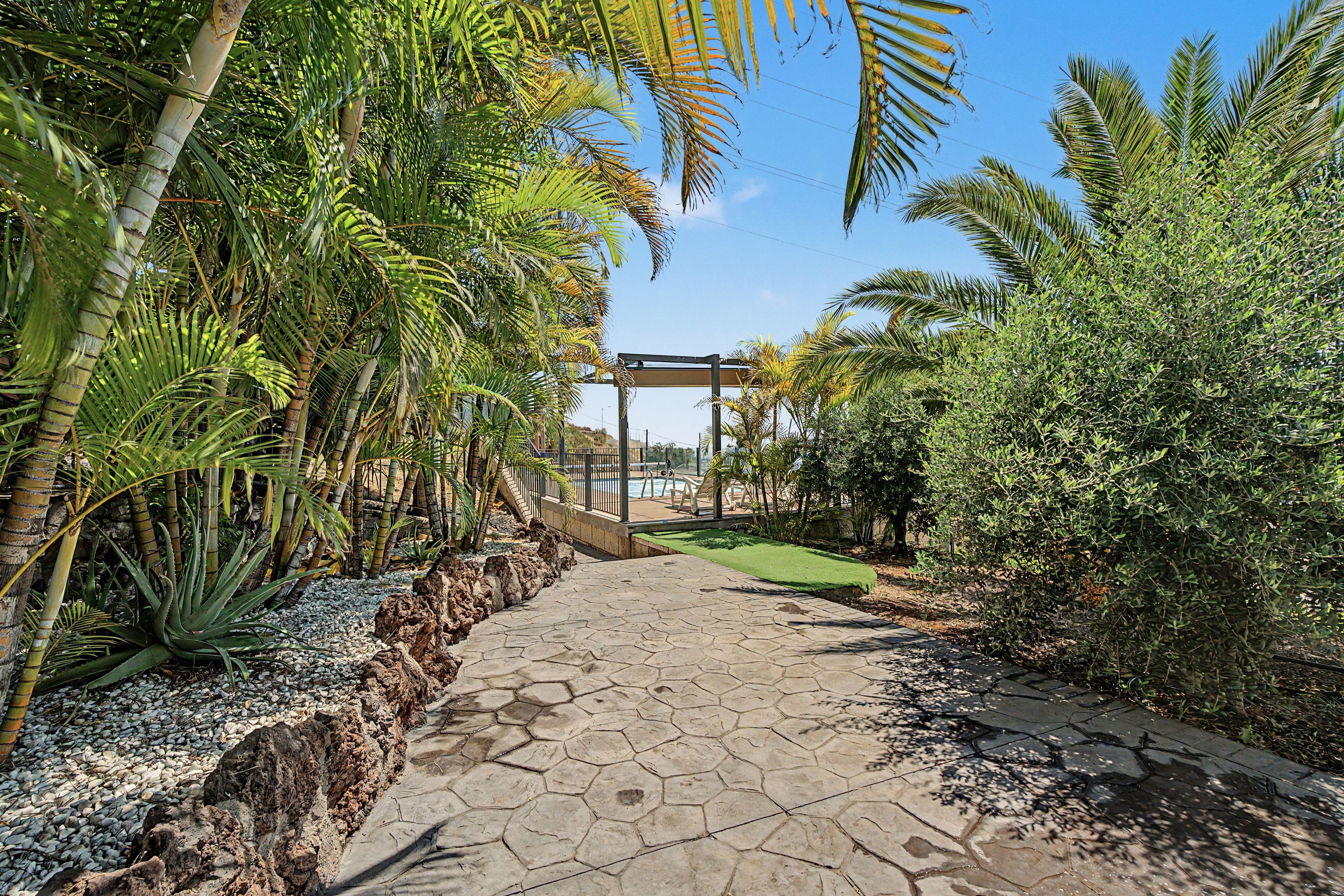 Un sentier pavé, bordé de végétation tropicale, mène à une zone abritée d’où l’on peut admirer une piscine.