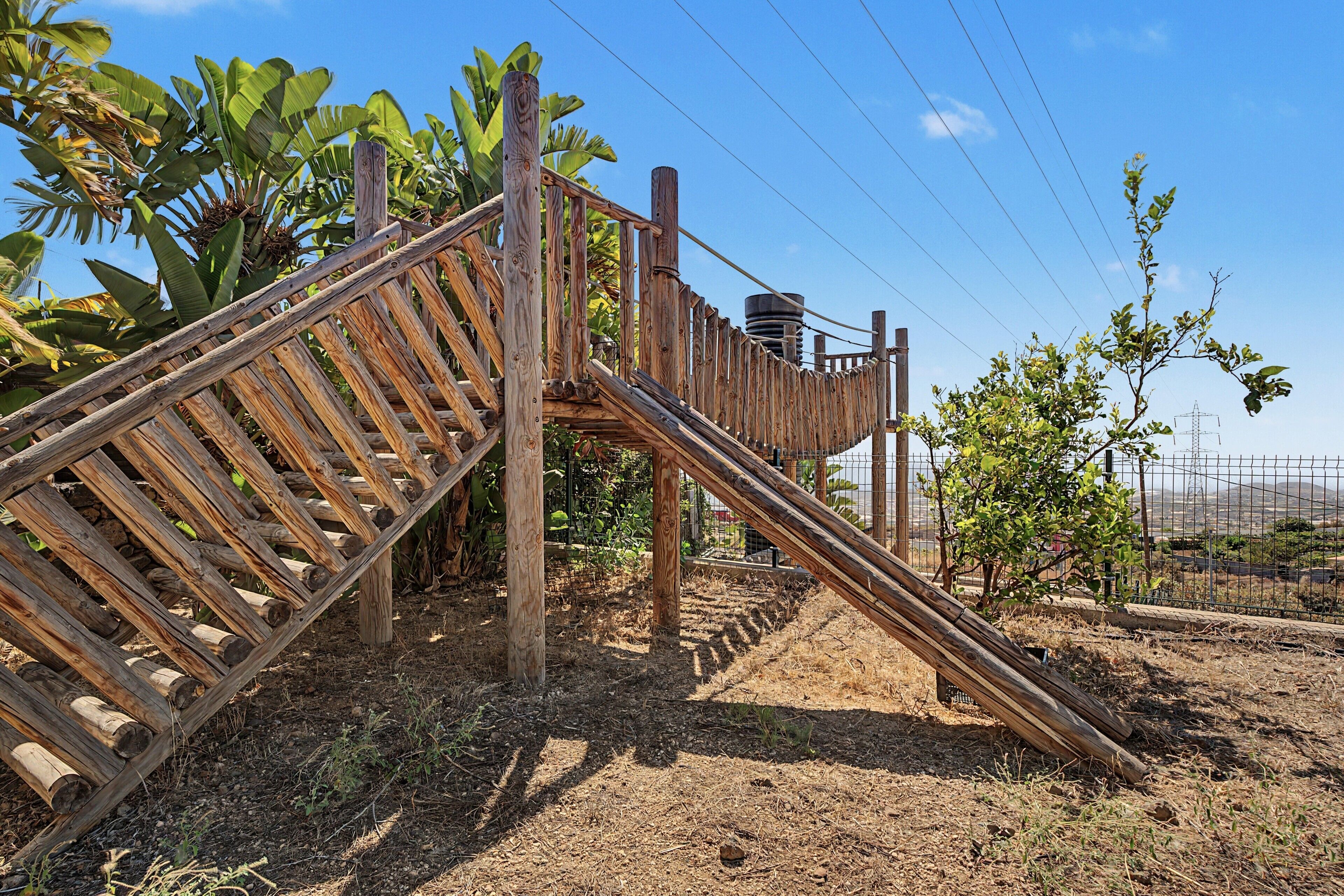 A wooden structure with a ladder and a slide, surrounded by dry grass and banana plants.