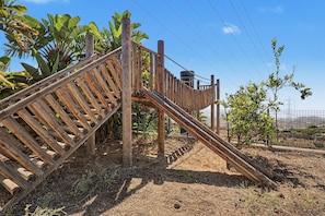Une structure en bois, dotée d’une échelle et d’un toboggan, entourée d’herbe sèche et de plants de bananiers.