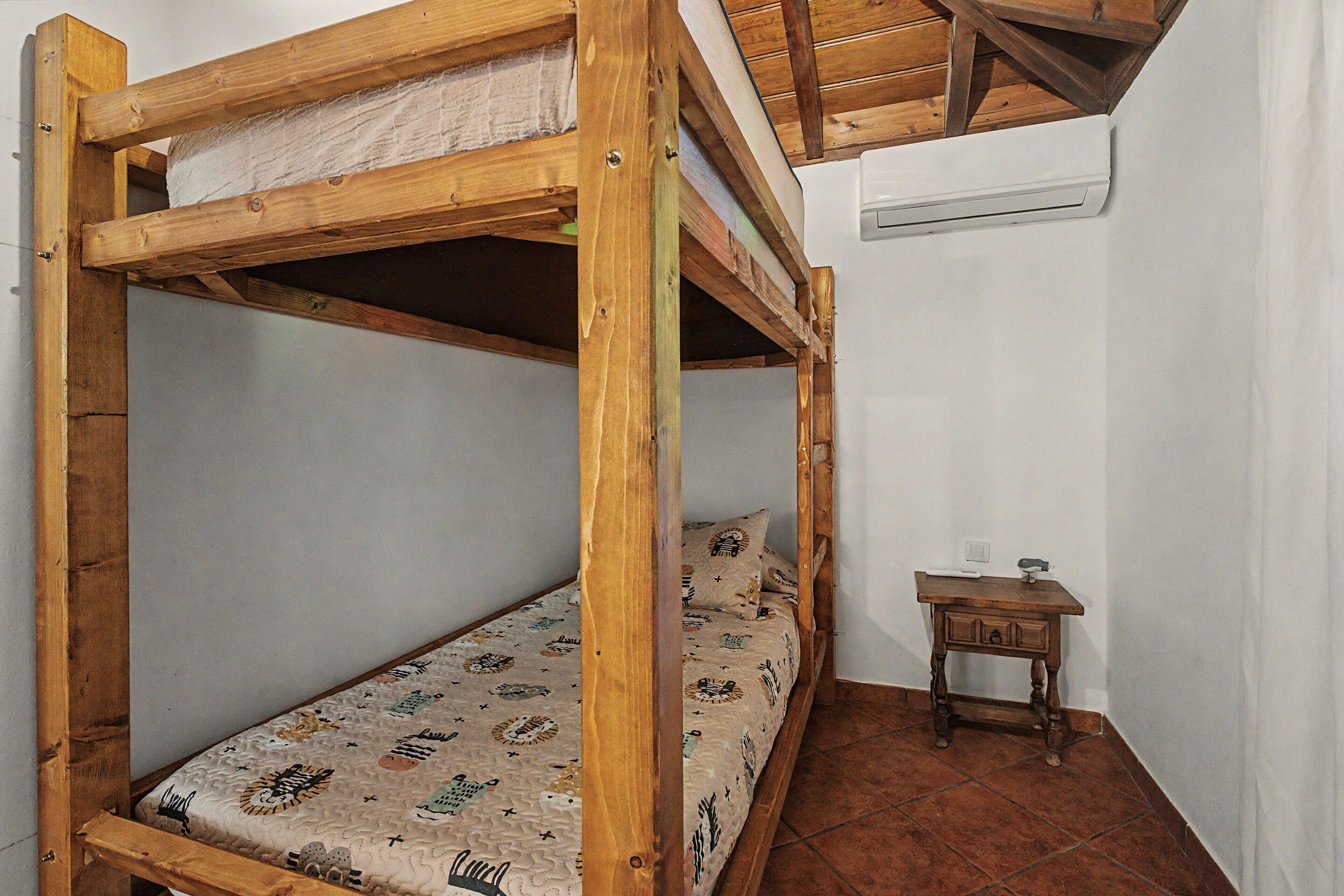 Una cama individual superpuesta con un colchón estampado, una mesita de noche de madera y una pared blanca con un termostato.