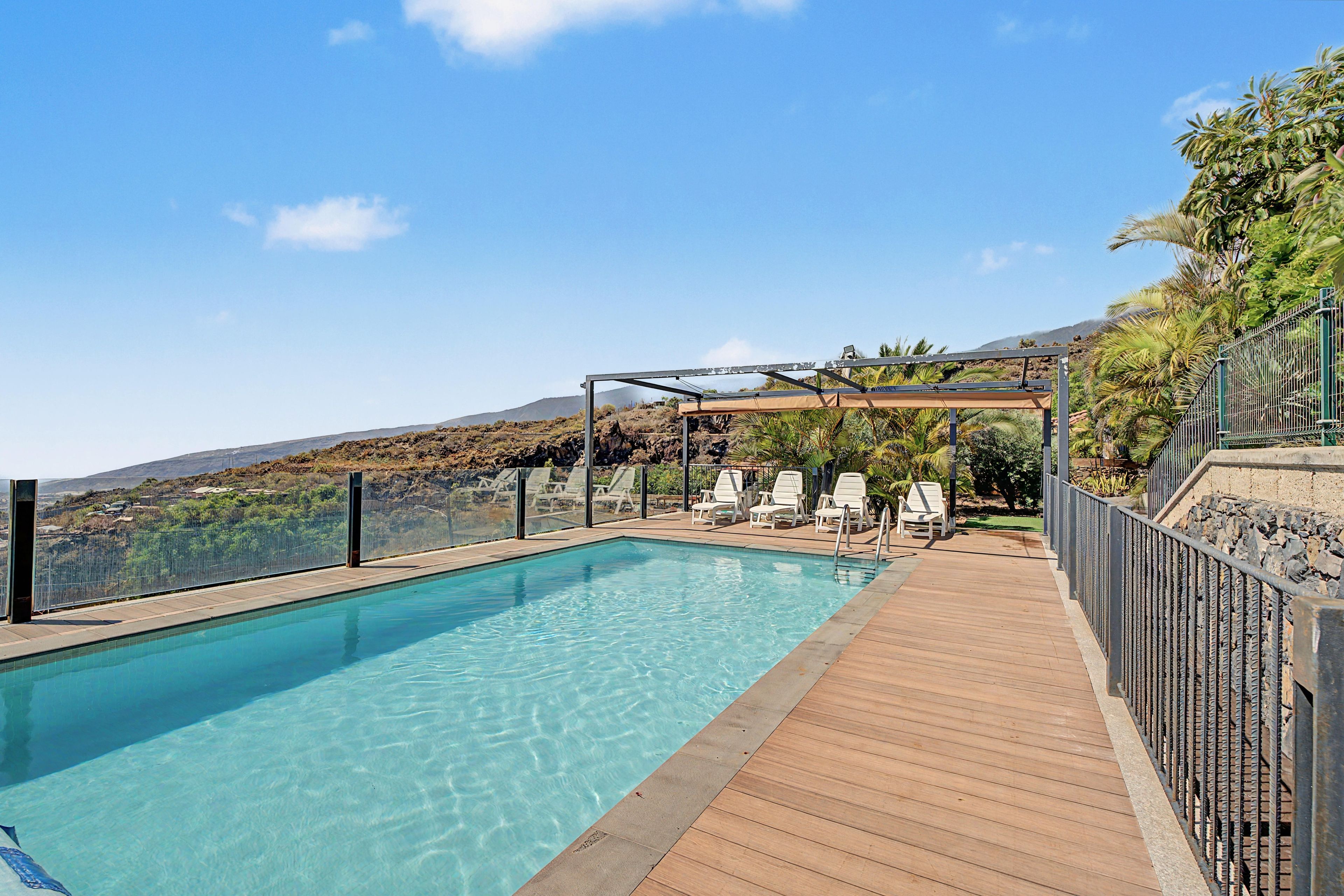 A clear swimming pool with a wooden deck, surrounded by a metal fence, and overlooking a hilly landscape.