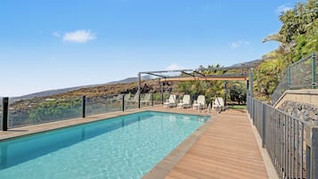 A clear swimming pool with a wooden deck, surrounded by a metal fence, and overlooking a hilly landscape.