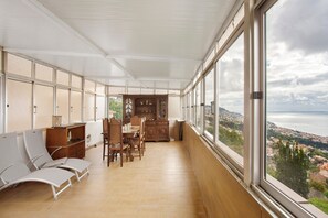 A room with a dining area, a seating area with chairs, and large windows offering a view of a coastal landscape.