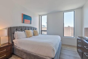 A bedroom with a large bed, a nightstand, a dresser, and two windows offering a city view.
