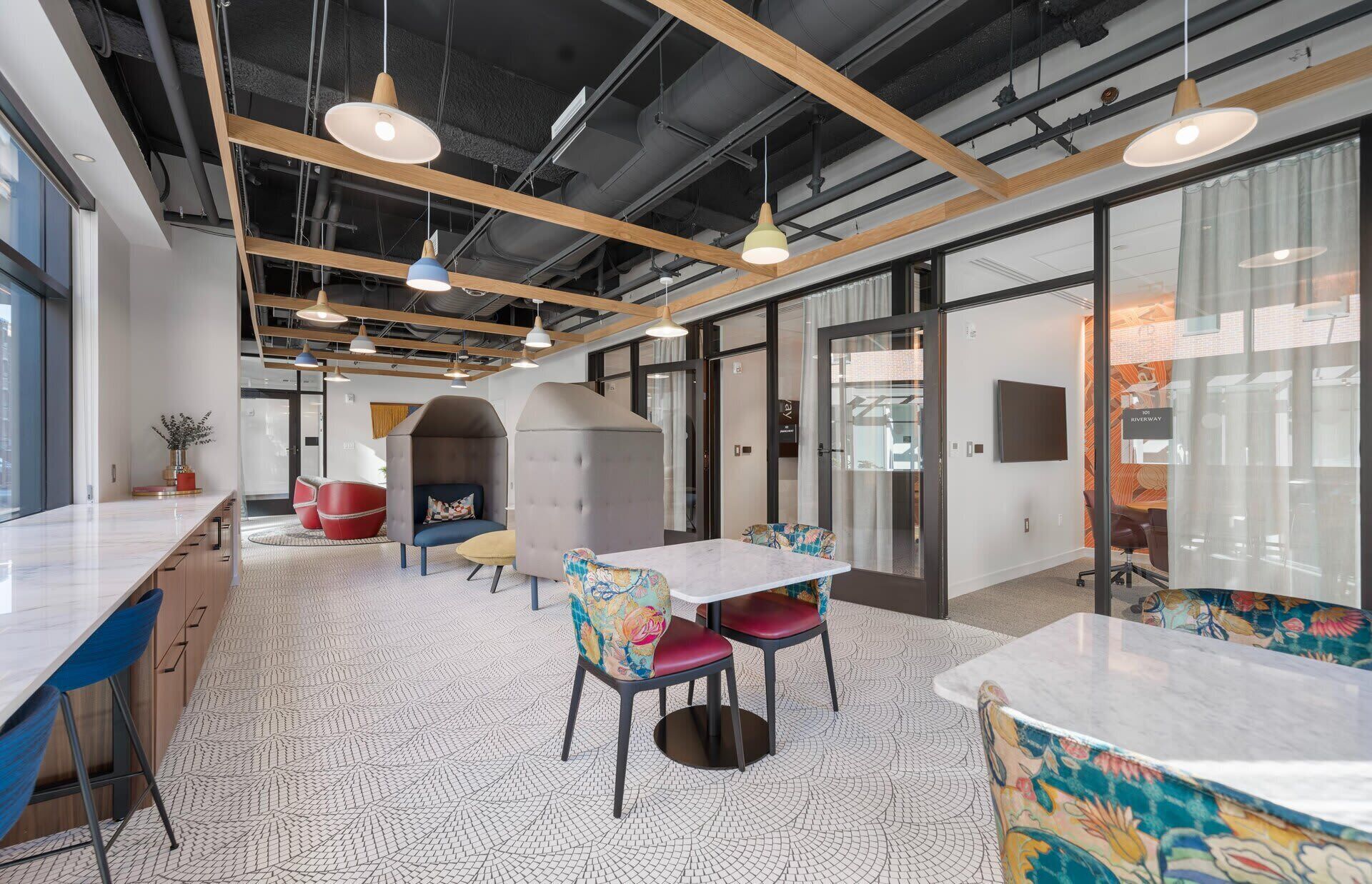 Un espace de bureau moderne, aménagé avec différents types de sièges, notamment une longue table et des chaises, un petit canapé et un fauteuil coloré. Des luminaires suspendus sont fixés au plafond, et la pièce est dotée d’un plafond haut avec des poutres apparentes.