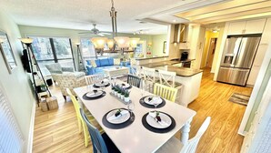 Une cuisine moderne dotée d’un îlot central, d’appareils électroménagers en acier inoxydable et d’un coin repas aménagé pour six personnes, avec une table blanche et des chaises bleues.