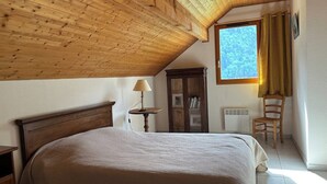 A bedroom with a wooden ceiling, a bed with a beige bedspread, a nightstand, a lamp, a chair, a wardrobe, and a window with curtains. - CHARMING CHALET - SLEEPS 6 (Allemont)