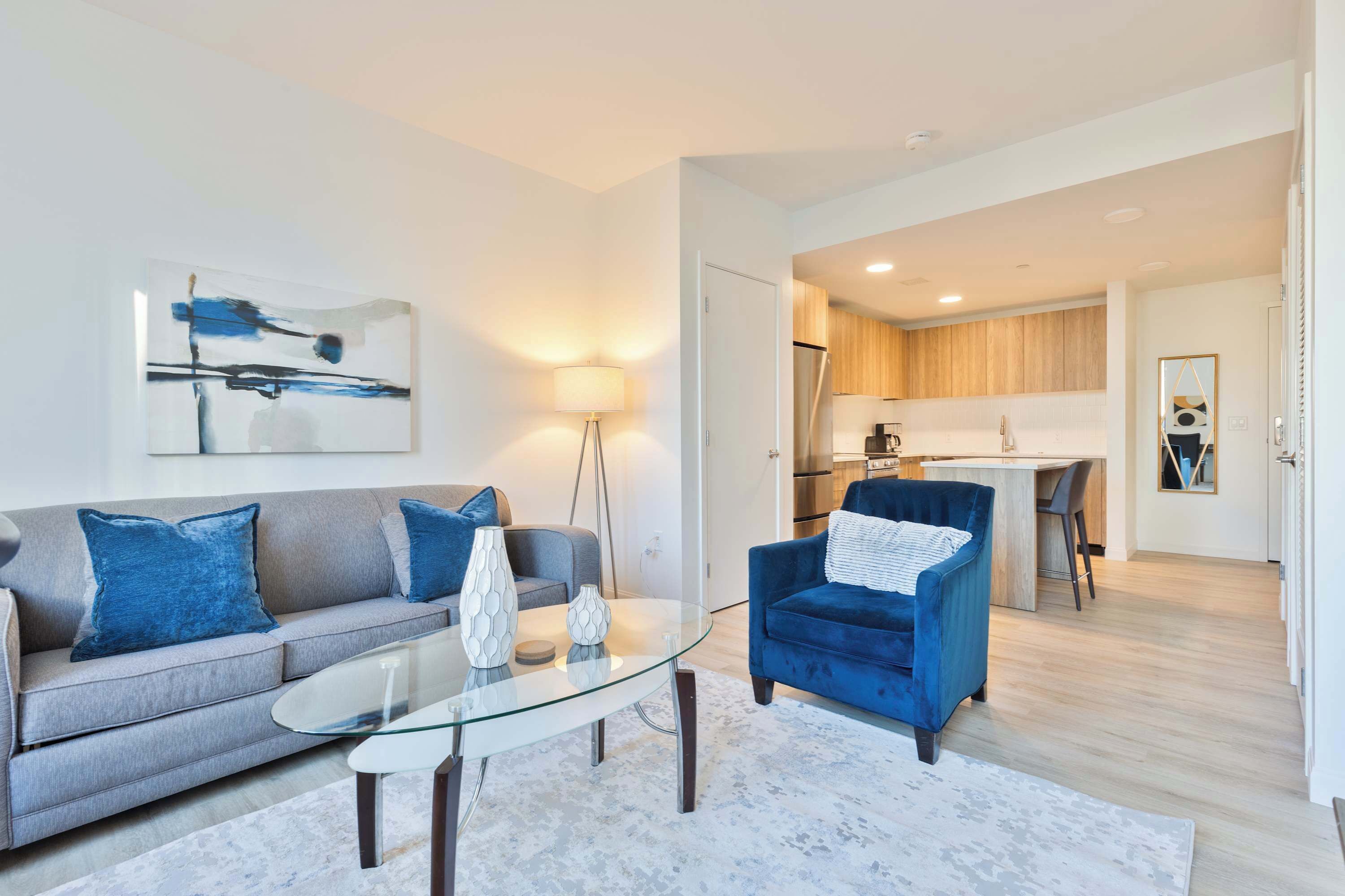 A modern living room with a glass coffee table, a sofa, and a blue armchair. A kitchen is visible in the background.