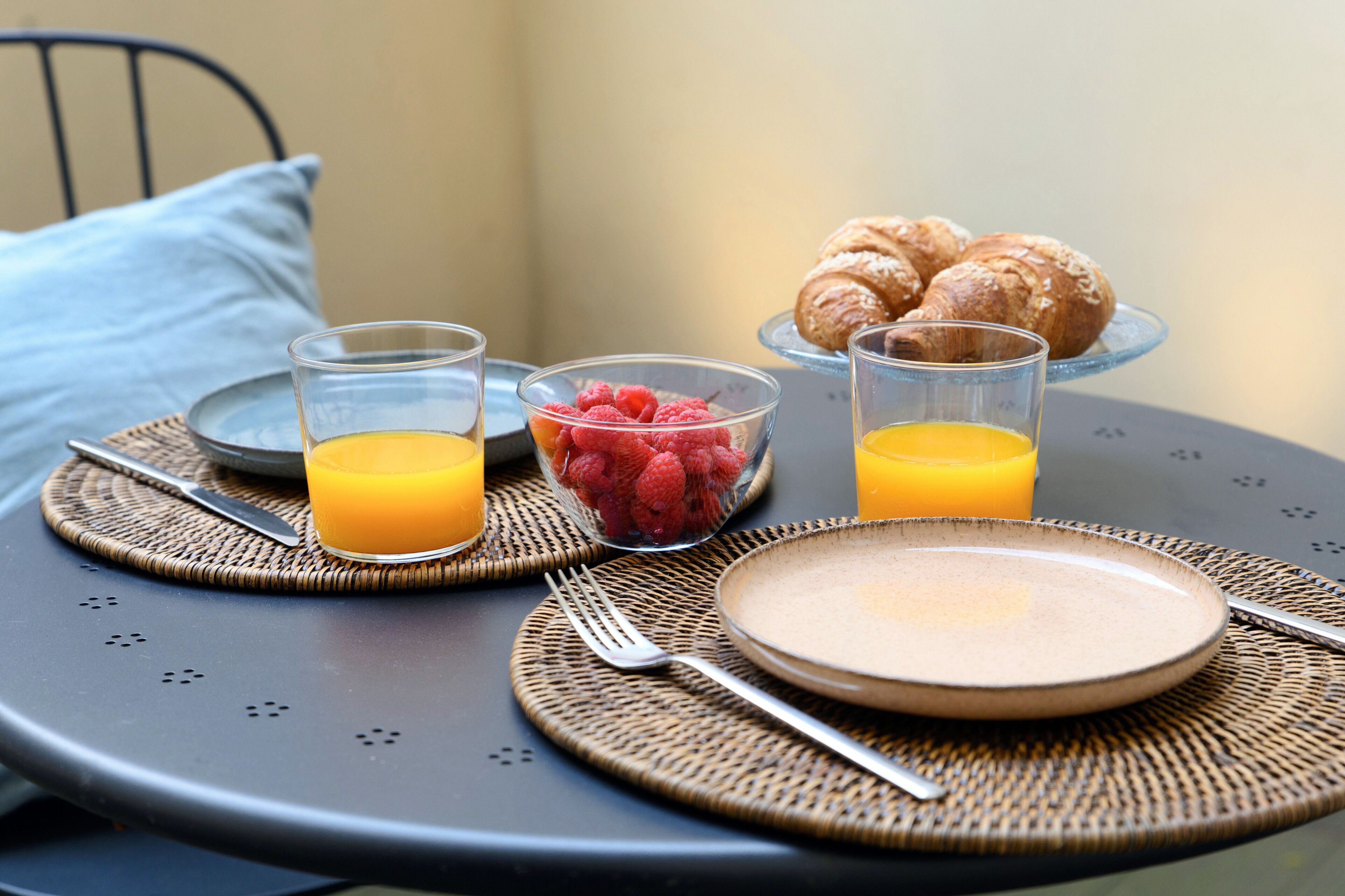 Desayuno con croissants, un tazón de frambuesas y vasos de jugo de naranja sobre una mesa redonda.